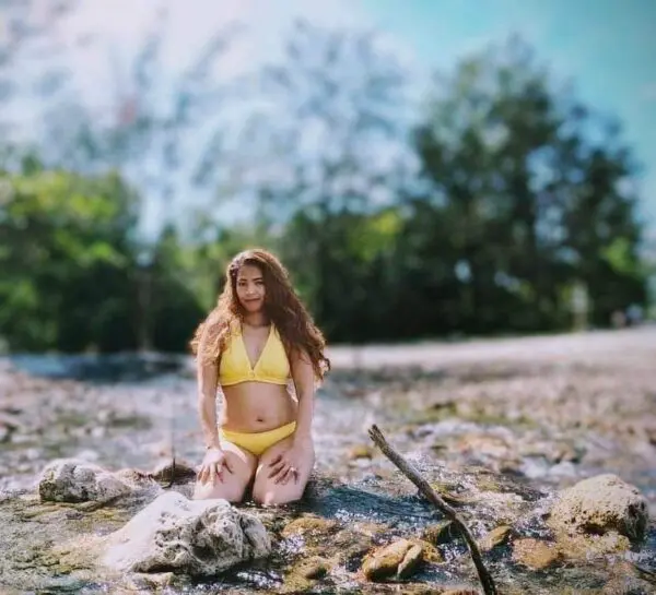 A woman in a yellow bikini poses on a rock in a river.