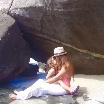A woman sitting on the beach under a large rock.